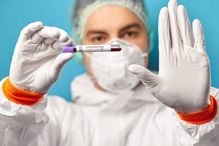 Doctor wears protective coverall and respiratory mask, holds medical test tube with a positive test...