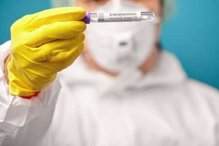 View of doctor in protective coveralls and respiratory mask, holding medical test tube with swab material taken from the mouth and nose to check for corona virus, close-up shot on a blue background.の写真素材