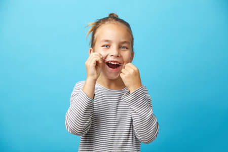 Portrait of smiling little five years old girl brushing her teeth with dental floss on blue isolated...