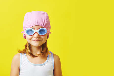 Time to swim. Funny little girl in swimsuit, cap, and glasses standing on yellow isolated background.の写真素材