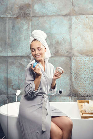 Woman in spa bathrobe sitting on bath, hold face cream and scrub in hand, smiling looks to camera.の写真素材