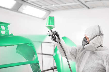 Male worker in protective clothes and mask painting car bumper using spray paint.
