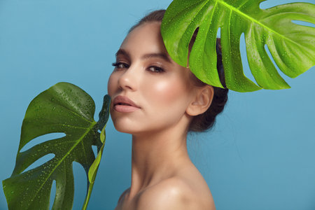 natural beauty. Attractive brunette lady with languishing look and perfect face near palm leaves on light blue background closeupの写真素材