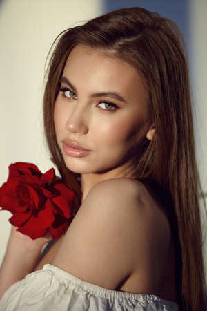 beauty and glamor. Portrait of gorgeous young woman with long dark hair in white dress holding fresh red rose on blurred backgroundの写真素材