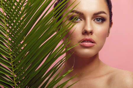 studio beauty portrait of beautiful caucasian brunette woman with branch of palm on pink isolatedの写真素材