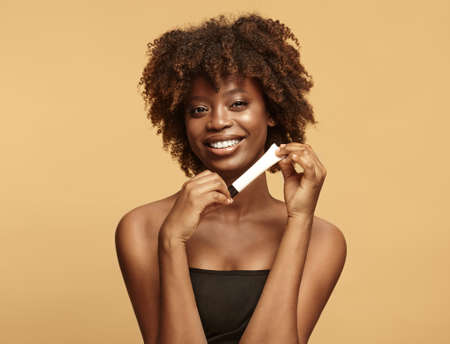 Smiling African American woman with a clean and healthy skin holding slim cream tube over beige...