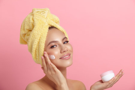 Smiling lady applies cream on face showing jar on pinkの写真素材