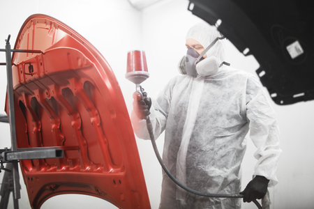 Automobile repairman painter in mask and protective workwear painting car hood in paint chamber.の写真素材