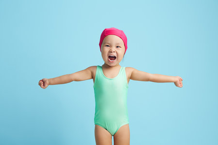 Little female child swimmer cheerfully spreads her arms to the sides, dressed in a turquoise swimsuit and a pink cap standing on a blue isolatedの写真素材