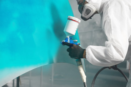 Professional spray painter in protective mask and clothes spraying car hood of vehicle part to blue color using spray gun in paint chamber.の写真素材