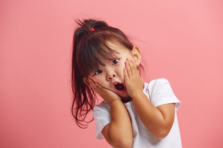 Headshot of stupefied little girl keeps hands on cheeks standing over pink isolated background....