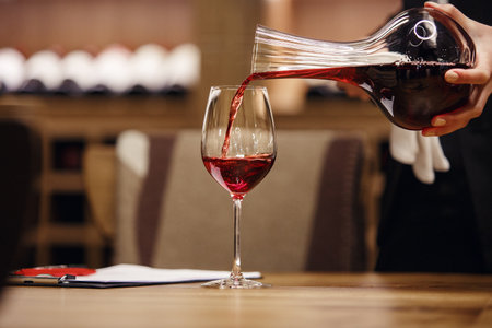 Professional female sommelier pours red wine from decanter to the glass, close up image. Woman waiter pouring alcoholic drink being in cellar of wine shop.の写真素材