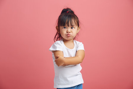 Offended little asian girl with insulted expression, scowls face, stick out lips in discontent, has a negative mood, dressed in white t shirt,crossed hands in front of her poses over pink background.の写真素材