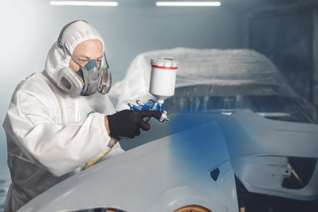 Car paint worker spraying blue paint to car body element using spray gun in vehicle workshop chamber. Complex restoration of automobile body.の写真素材