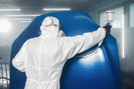 Professional spray painter in protective mask and clothes spraying car hood of vehicle part to blue color using spray gun in paint chamber.の写真素材