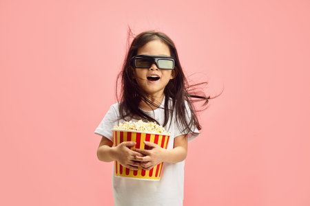 Joyful and Surprised Child Girl in 3D Glasses Holding Popcorn Bucket Having Flowing Hair Standing...