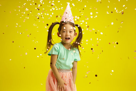 Cheerful Asian Child Girl Celebrating Birthday with Confetti on Yellow Isolated Background