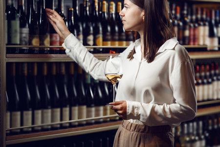Female sommelier holding glass with white wine choose bottle in cellar.の写真素材