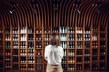 Female sommelier stands in wine cellar with large shelves full of closed bottles with exquisite alcohol drinks put in big collection.の写真素材