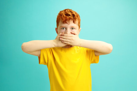 Little Red Headed Boy Covers Mouth With Hands, Looking to Camera, doesnt Want Say Anything, Wearing in Yellow T-shirt Standing Over Blue Isolatedの写真素材