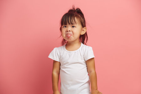 Little Asian baby girl shows tongue wears t shirt isolated over pink background. Three years old offended and capricious child.の写真素材