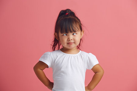 Funny little asian girl crosses eyes, foolishness indoor, has fun, dressed in t shirt isolated over...