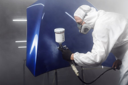 Worker in protective mask and jumpsuit overalls spraying finishing coat of varnish on car body...