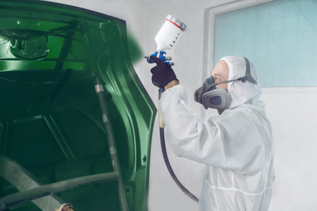 Worker in protective mask and jumpsuit overall spraying base coat of paint on car body elements in painted chamber using professional airbrush.の写真素材