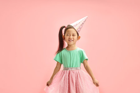 Beaming Child Girl in a Pink Tutu, Festive Turquoise T Shirt and ice cream Cone Party Hat Celebrates Herself Birthday Joyfully Standing on Pink Isolated Background.の写真素材