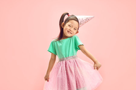 Cheerful Birthday Asian Child Girl in Festive Clothes, Having Party Hat on Head, Cute Raising Shoulders, Funny Straightens her full Skirt to the Sides Standing Over Pink Isolated Backgound.の写真素材