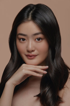Vertical close up portrait of beautiful asian female model with a healthy clean skin touching her face on a beige background.の写真素材