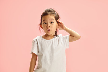amazed little girl has curiosity putting hand to ear and overhear something expresses surprise, cant believe what she heard standing on pink isolated backgroundの写真素材