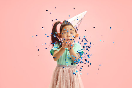 Waist up portrait of Asian Child Girl Blowing Blue Glitter to Camera While Standing Against Pink Isolated Backgroundの写真素材