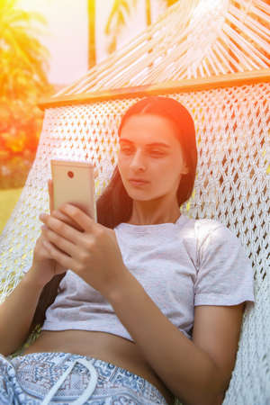 Relaxed young woman looking at mobile phone in hammock. Tonedの写真素材