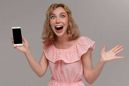 Portrait of a happy cute girl in dress pointing at mobile phone and looking at camera isolated over gray backgroundの写真素材