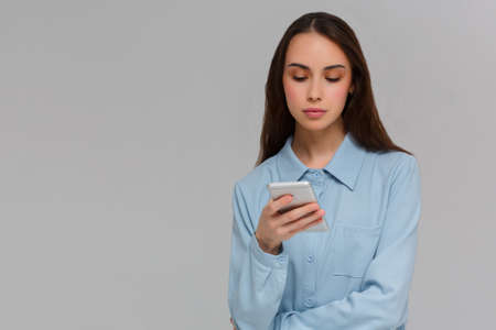Content teenager with long hair, holds modern cell phone, scrolls through social networks, has cheerful expression, isolated over gray background, recieves messageの写真素材