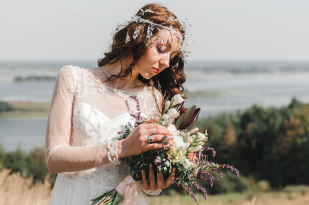 The bride in a white dress with a beautiful bouquet of fresh flowers, standing on a hill with views of the river flood.の写真素材