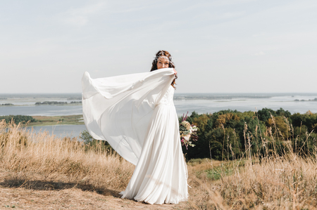 The bride in a white dress with a beautiful bouquet of fresh flowers, standing on a hill with views of the river flood.の写真素材