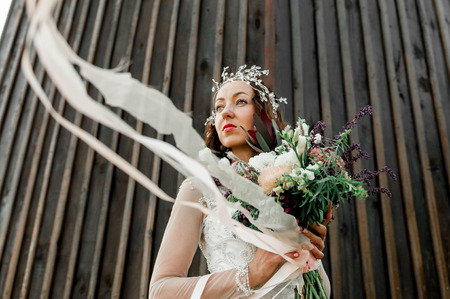 The bride with a beautiful green background bouquet standing on a dark wall with vertical lines, smiles and ribbons on the bouquet develops in the windの写真素材
