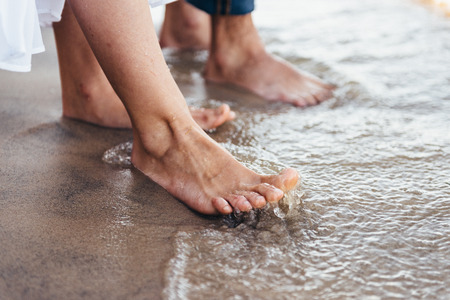 a man and a woman stand barefoot on the wet sand, their gogi is washed by water with foam and sprays. girl in white dress hanging down to waterの写真素材