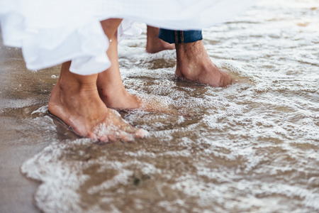 a man and a woman stand barefoot on the wet sand, their gogi is washed by water with foam and...