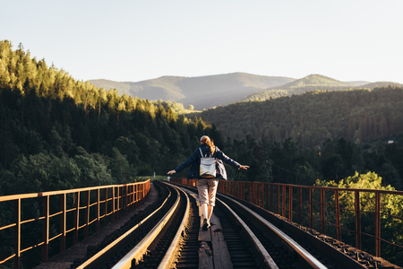 A tourist girl in the mountains on a railway bridge with her hands apart in the form of a bird, on a background of high mountains and their peaks, in summer at sunsetの写真素材