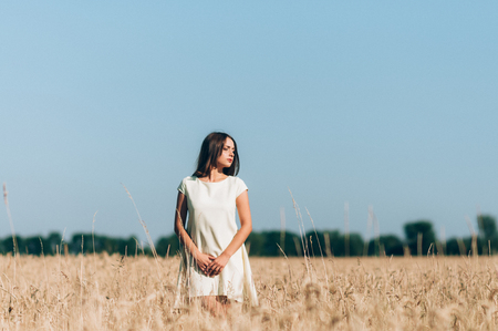A girl in a white dress in the middle of a wheat field in a sunny summer day against a blue sky and a forest lineの写真素材