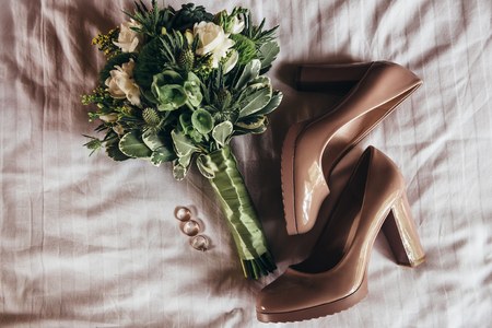 a lively green wedding bouquet with white and yellow flowers tied with a green ribbon, lies on a sheet next to the peach-colored bride's shoes and wedding engagement ringsの写真素材