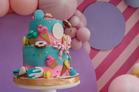 Bright colored handmade cake with edible, sweet decorative elements and macaroons for a children's party, against a pink wall with balloonsの写真素材