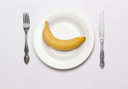 One banana on white plate with fork and knife on white background. Concept image for diet.の写真素材