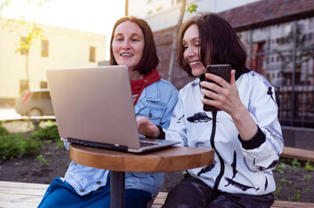 Two smiling young women using laptop and phone at the park, laughing, chattingの写真素材