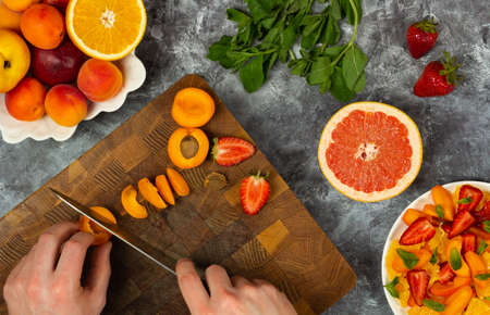 Chopping fruit salad on wooden cutting board. Healthy tasty food on dark concrete background. Ingredients for fruit salad. From above, flat, overhead.の写真素材