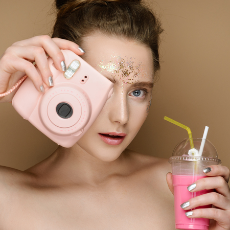 Portrait of young beautiful girl with pink milk shake coctail and camera in hands on the gold background. Nude makeup with tinsel and updo. Concept of the beauty.の写真素材