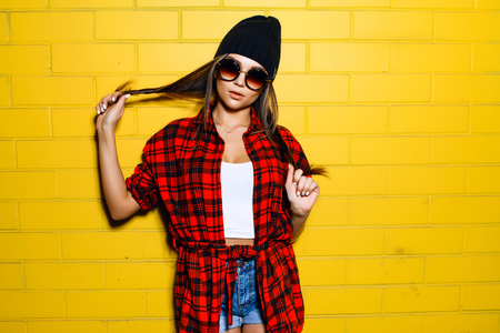 Beautiful young sexy hipster girl posing and smiling near urban yellow wall background in sunglasses, red plaid shirt, shorts, hat.の写真素材